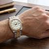 An image of a pre-owned men's Rolex wristwatch displayed on a wrist. The watch is positioned at a slight angle, showing the face and part of the band. It features a date indicator and luminous hands. The band is a combination of gold and stainless steel tones. The image is taken from a close distance, capturing the details of the watch against a wooden table background with a pen and notebook nearby.