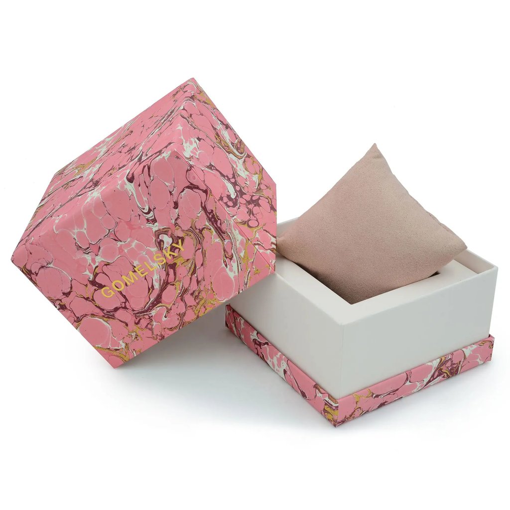 An image of a wristwatch box taken from a slightly elevated angle, showcasing the open packaging. The box is predominantly pink with an abstract marbled design and the brand name "GOMELSKY" in gold lettering. The lid is positioned to the left, partially covering the bottom half, which contains a beige, wedge-shaped pillow intended to hold the watch. The image is captured at a close distance, highlighting the box's texture and design details.