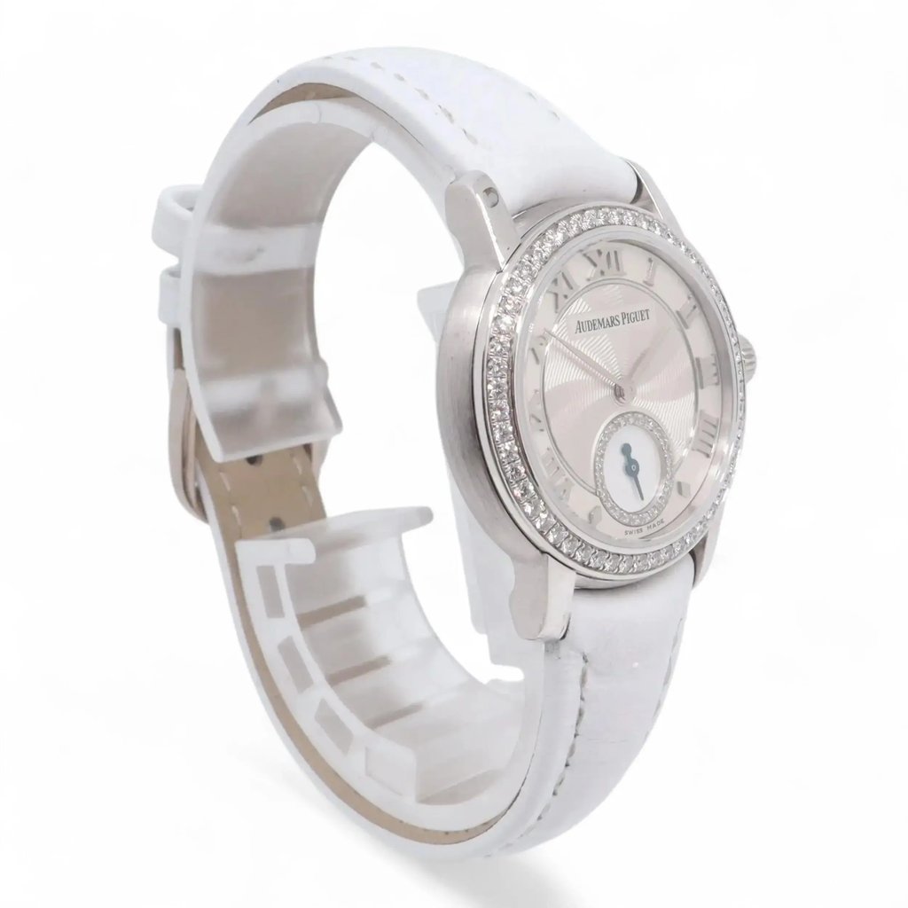 An image of a pre-owned Audemars Piguet women's wristwatch. The watch is positioned at a slight angle, showcasing the face and part of the strap. The silver watch face features Roman numerals and a small subdial, encircled by a band of sparkling diamonds. The white strap is partially visible, with the watch standing upright and slightly turned to the right. The photo is taken from a close distance, capturing the intricate details of the watch design.
