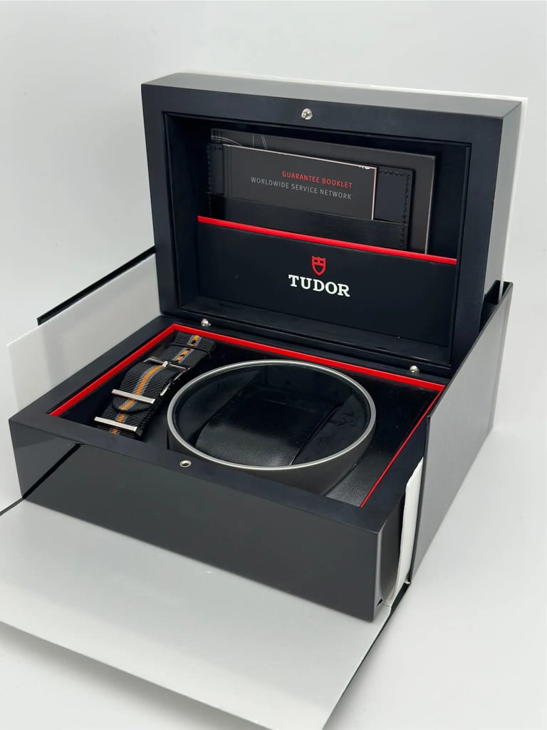 An image of an open wristwatch box positioned at a slight angle, viewed from a close distance. The black box features the brand name "Tudor" on the inside lid, with a red interior trim. A guarantee booklet is visible in a pocket on the lid. The watch cushion is empty, and a striped strap is placed next to it.