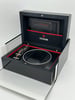 An image of an open wristwatch box positioned at a slight angle, viewed from a close distance. The black box features the brand name "Tudor" on the inside lid, with a red interior trim. A guarantee booklet is visible in a pocket on the lid. The watch cushion is empty, and a striped strap is placed next to it.
