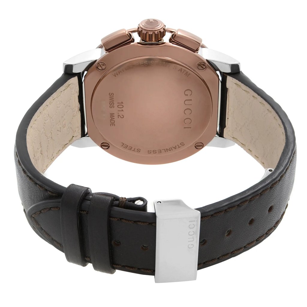 An image of a pre-owned Gucci men's wristwatch viewed from the back, showing a stainless steel caseback and leather band. The watch is positioned at a slight angle where the top of the watch faces slightly to the left with the deployant clasp partially visible and closed. The round caseback is solid with the brand name and other details engraved, and the leather strap is in the foreground, curving from the bottom of the image towards the upper right corner, suggesting the watch is laid flat. The band exhibits a dark color with stitching details, and a price tag is attached near the clasp. The distance from the camera to the watch allows for a clear and detailed view of the textures and inscriptions on the caseback and strap.