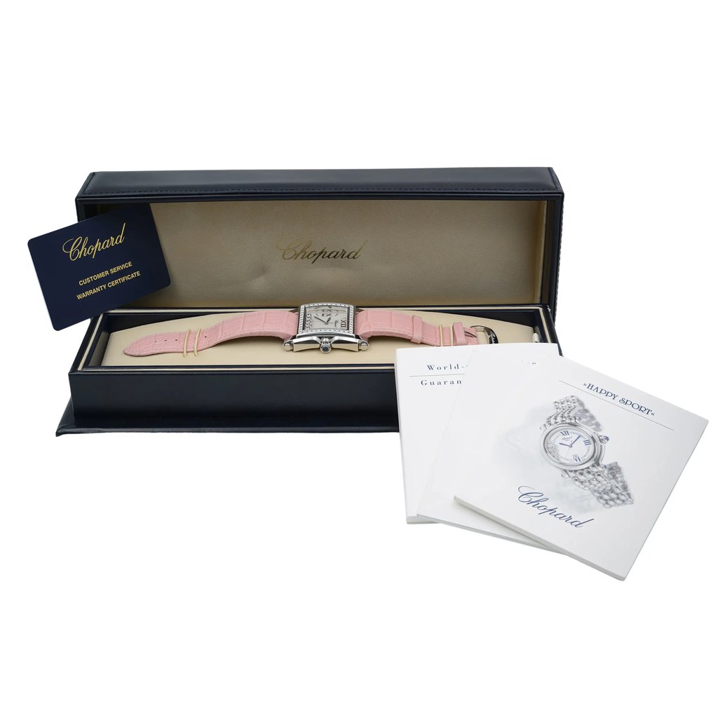 An image of a pre-owned wristwatch positioned inside an open watch box. The watch features a pink strap and is centered within the box, which is displayed from a slightly elevated angle. The box is navy blue with a beige interior, and the brand name is visible inside the lid. To the right of the box, there are two documents partially overlapping, one labeled "World" and the other "Happy Sport." A customer service and warranty certificate card is positioned leaning against the open lid on the left side. The shot is taken at a medium distance, capturing the full view of the box, watch, and accompanying documents.