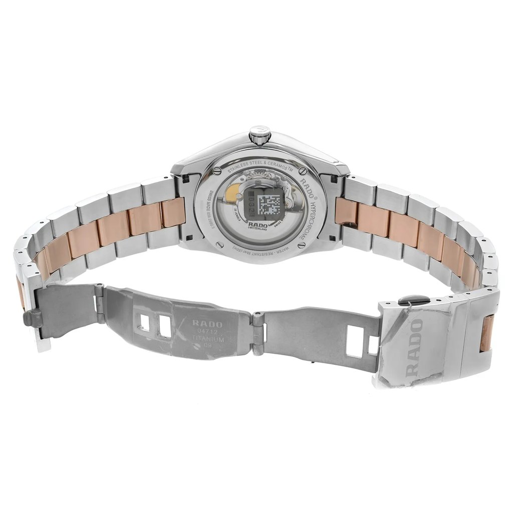 An image of a wristwatch taken from the back view, showing the open clasp. The watch is a display model, with a clear view of the back case, revealing internal mechanisms. The watch has a metal strap with alternating silver and rose gold links, positioned centrally, at eye level, and at a moderate distance, highlighting the craftsmanship of the clasp and bracelet.