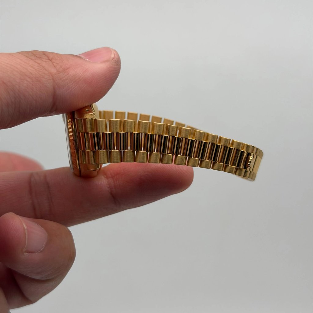 An image of a pre-owned women's Rolex wristwatch held between two fingers. The focus is on the gold bracelet, viewed from the side at a close distance, showcasing its metallic links and its stretch.