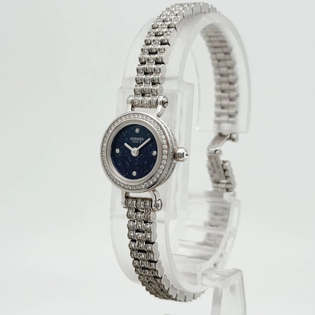 An image of a pre-owned women's HERMÈS wristwatch positioned upright on a clear stand. The watch is angled slightly to the right, showcasing its intricate white gold link bracelet and a round, dark blue watch face adorned with diamond markers. The distance provides a clear view of the elegant design and craftsmanship.