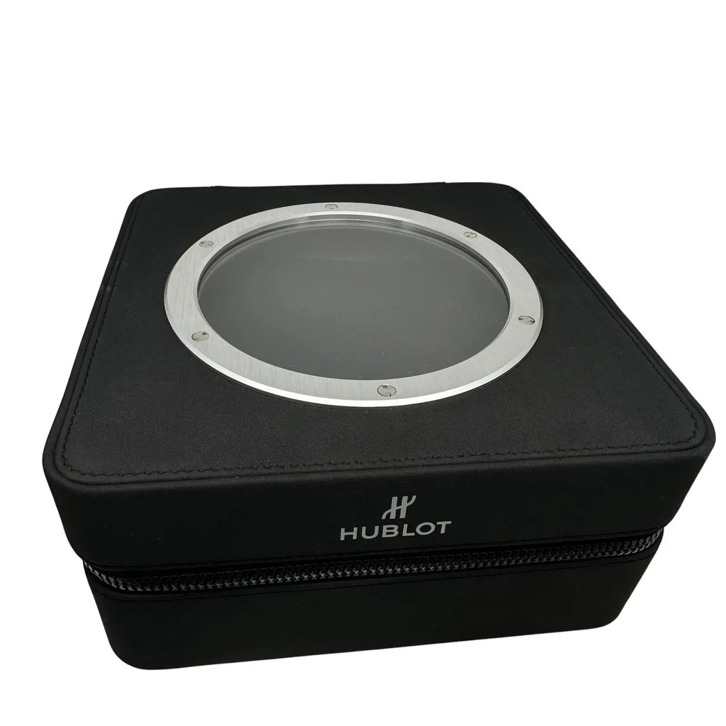 An image of a Hublot wristwatch box taken from a slightly elevated angle, showcasing the full front view. The box is black with a circular, transparent window on top, framed by a metallic ring with screws. The brand name "Hublot" is visible on the front side. The image is taken at a close distance, focusing on the details of the box.