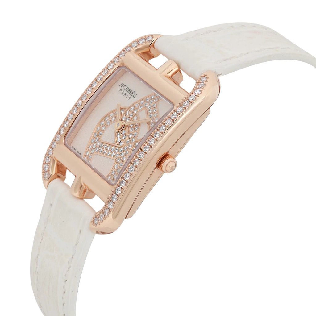 An image of a pre-owned women's Hermès Cod Joaillier wristwatch taken at a slight angle from the front and side. The watch features a rectangular face with a 18k rose gold case, encrusted with diamonds along the bezel. It  has a white leather strap attached to the case. The watch's elegant design includes intricate detailing on the face, visible at a close distance.