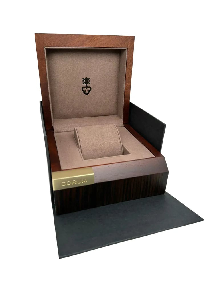 An image of an open wristwatch box taken from a slightly elevated front angle. The box is empty, showcasing a plush interior with a cushion for the watch. The lid is propped open, revealing a subtle emblem on the inner side. The outer edges of the box are dark wood with a metallic nameplate on the front edge. The image is captured from a medium distance, highlighting the box's craftsmanship and materials.
