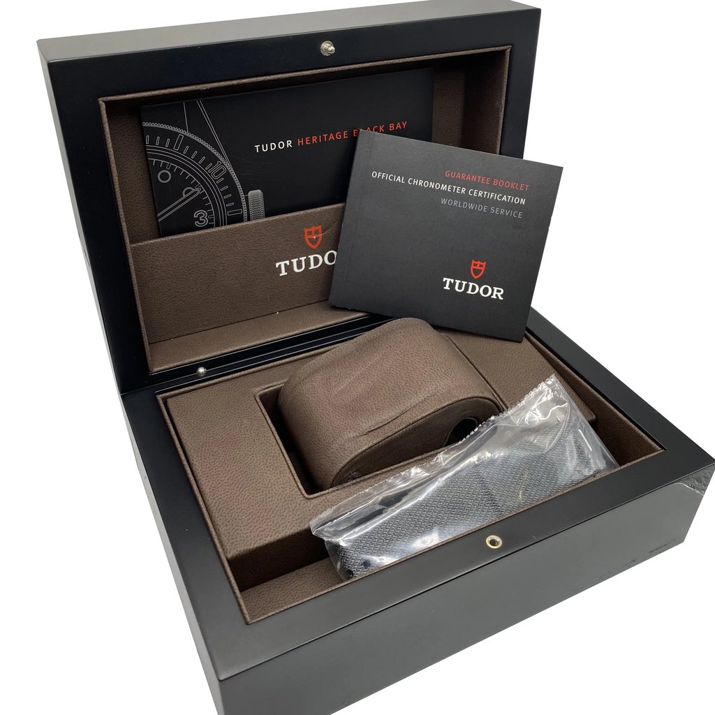 An image of a wristwatch box set viewed from a slightly elevated angle. The image shows an open black box containing a brown cushion and a plastic-wrapped strap, with no watch present. Two booklets are visible inside the box, one titled "Tudor Heritage Black Bay" and the other "Official Chronometer Certification." The image captures the box's interior details from a close distance.