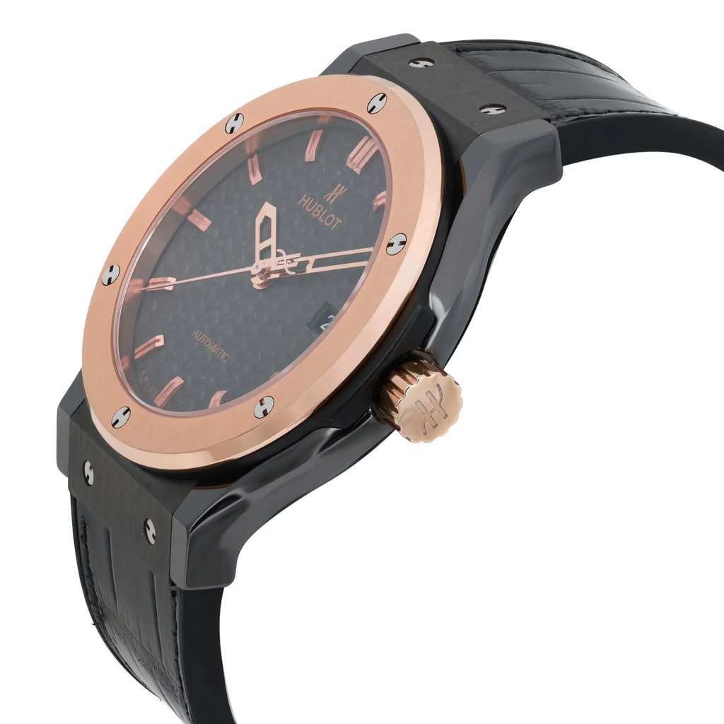 An image of a pre-owned Hublot men's wristwatch, positioned at a slight angle from the left. The watch features a black strap and a rose gold bezel. The face of the watch displays a date indicator and rose gold hour markers. The crown is prominently visible, showcasing the brand's logo. The image is taken from a close distance, highlighting the watch's details and craftsmanship.