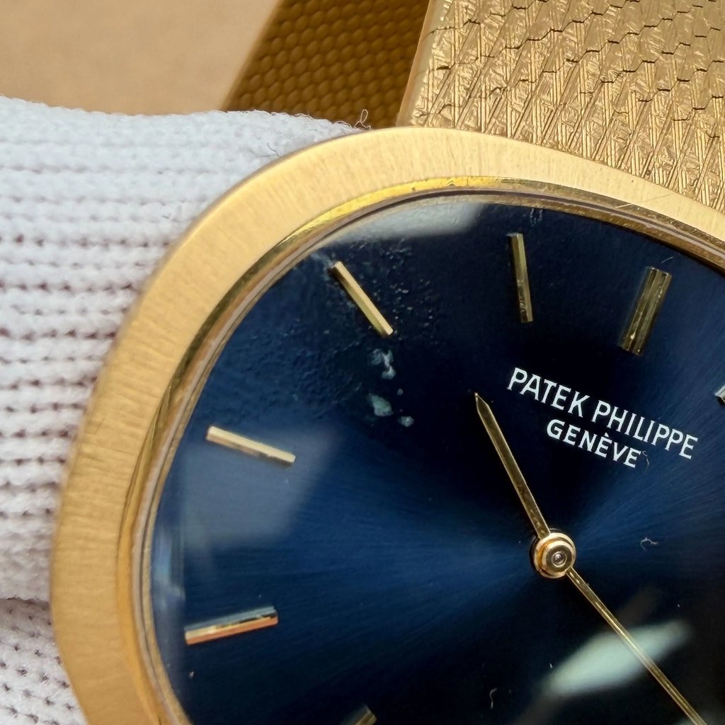 An image of a pre-owned Patek Philippe unisex wristwatch, captured close-up from an angled side view. The watch features a gold-toned case with a textured band and a deep blue dial with gold hour markers and hands. The brand name "Patek Philippe Genève" is visible on the dial. The watch is held in a white gloved hand, highlighting its luxurious design.