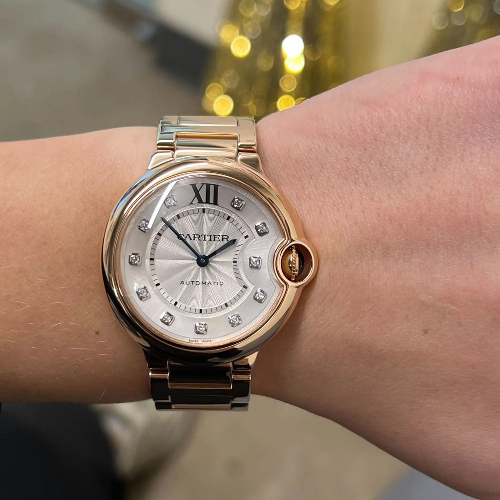 An image of a pre-owned Cartier luxury wristwatch for women, presented on a wrist at close range. The watch has a round rose gold case and band, featuring a silver dial with Roman numerals at the 12 o'clock position and diamond markers. The watch is viewed from a direct angle, with the dial fully visible and the band partially wrapped around the wrist. The photo is taken from a close distance, emphasizing the watch's details and craftsmanship.
