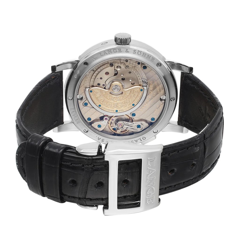 An image of a pre-owned wristwatch showcasing its back view. The angle focuses on the intricate mechanics of the watch's movement, highlighting the visible gears and components through a transparent case back. The watch is positioned upright, with the black leather strap and metal clasp prominently displayed in the foreground. The clasp is engraved with the brand name "LANGE." The photo is taken from a close distance to capture the fine details of the craftsmanship.