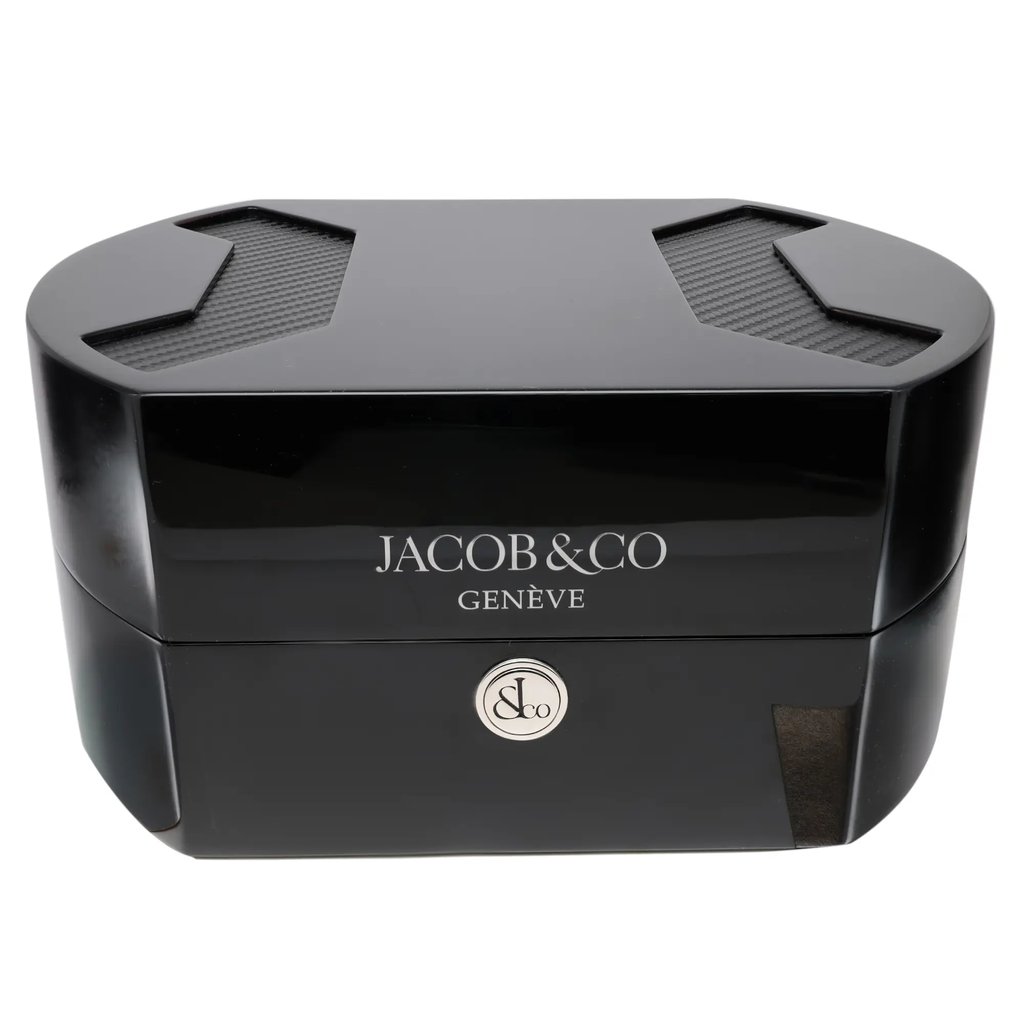 An image of a black wristwatch box from Jacob & Co Genève. The box is positioned centrally in the frame, viewed from the front at eye level. It features a glossy finish with the brand logo prominently displayed on the front, and a circular emblem below it. The top of the box has two textured, recessed areas, adding a touch of elegance.
