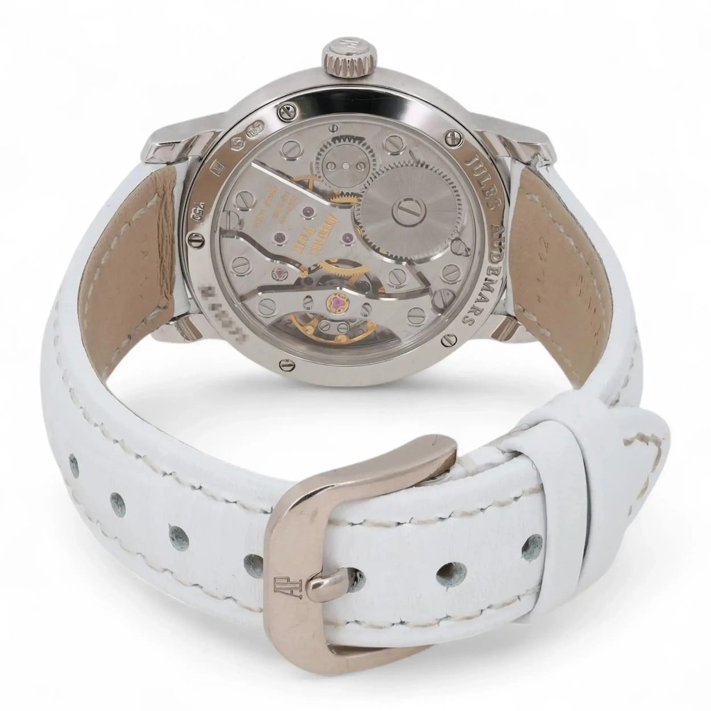 An image of a pre-owned Audemars Piguet women's wristwatch, viewed from the back at a slight angle. The focus is on the transparent case back, showcasing the intricate mechanical movement inside. The watch features a white leather strap with visible stitching, and the metal buckle is prominently displayed in the foreground.