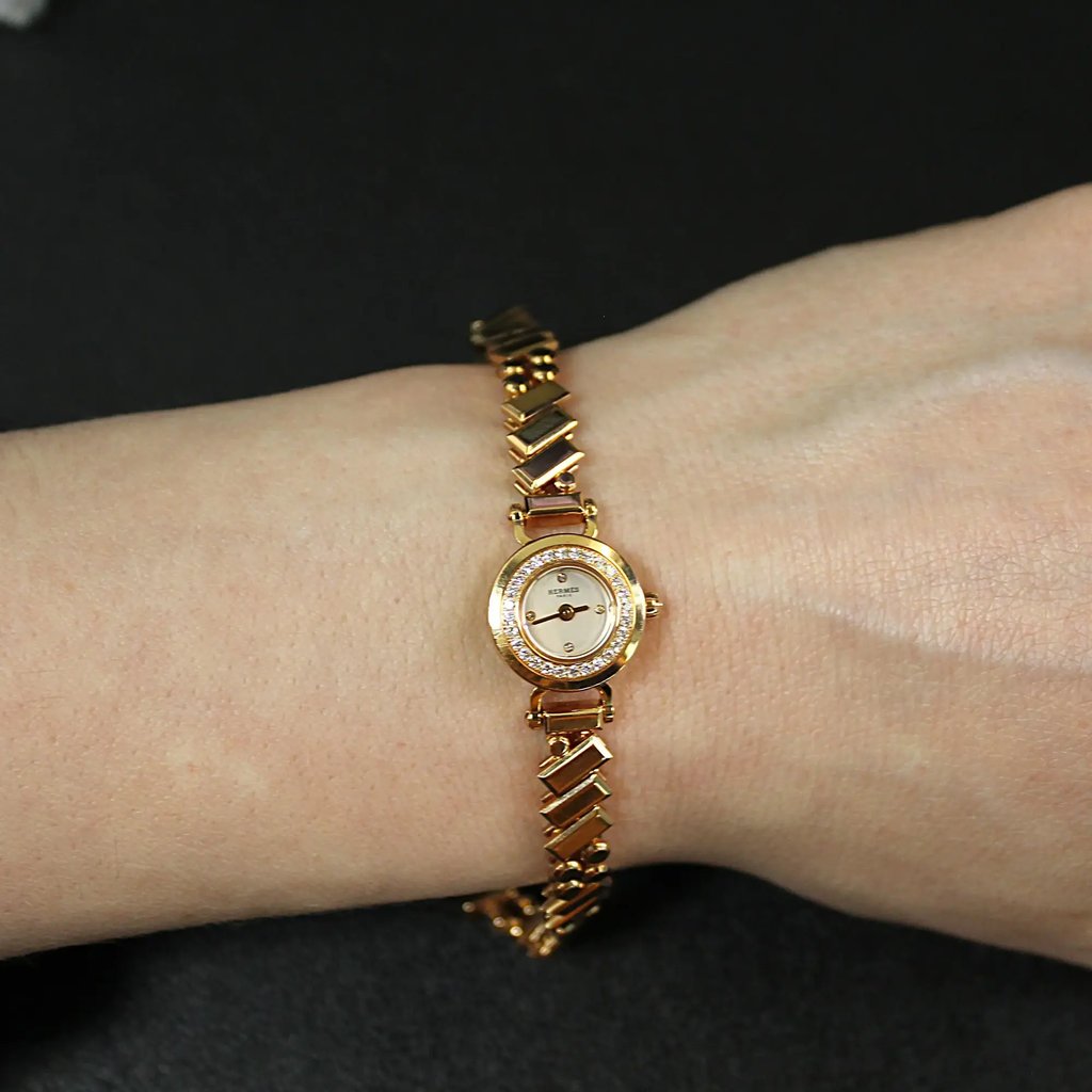 An image of a pre-owned women's HERMÈS wristwatch displayed on a wrist. The watch features a rose gold bracelet and a small, round face with a diamond-studded bezel. The watch is positioned centrally in the image, taken at a close-up angle from above, showcasing the elegant design against a dark background.