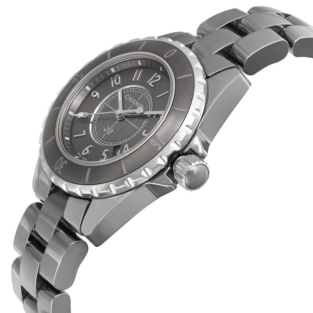 An image of a pre-owned CHANEL wristwatch in a side profile view, displaying the crown on the right side. The watch has a round shape with a gray dial and bezel, and features such as a date indicator and luminous hands. The ceramic band and case exhibit light scratches and signs of wear, indicating regular use. The photo is taken from a close distance, focusing on the side angle to highlight the watch's silhouette and the texture of the ceramic materials.