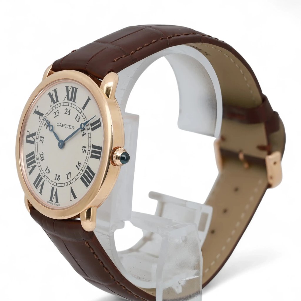 An image of a pre-owned Cartier unisex wristwatch positioned at a slight angle on a clear stand. The watch features a round face with Roman numerals and a dark brown leather strap. The photograph is taken from a close distance, highlighting the sleek design and gold casing.