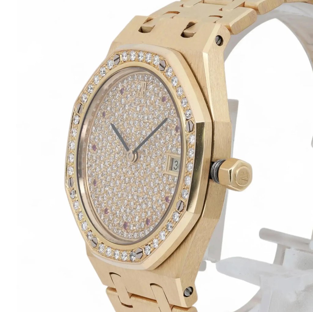 An image of a pre-owned Audemars Piguet women's wristwatch, viewed from a slightly angled side perspective. The watch features a gold bracelet and a diamond-encrusted bezel. The face is adorned with numerous small diamonds, with black hands and a date indicator visible on the right side. The crown is positioned prominently on the right, showcasing intricate detailing. The watch is displayed on a stand against a neutral background.