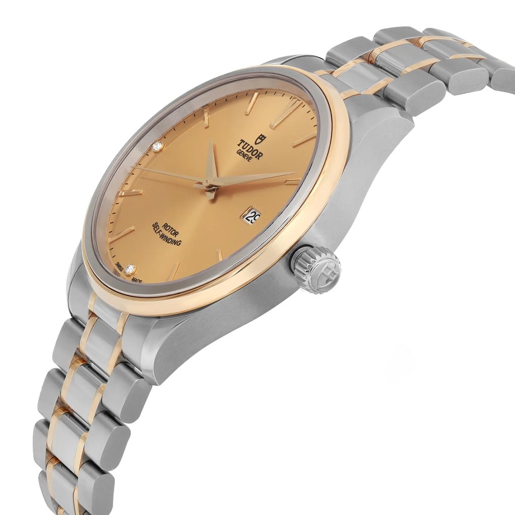 An image of a wristwatch shown from a side profile view, highlighting the crown. The watch features a gold-tone face with minimalistic hour markers and a date window. The casing and bracelet are crafted from a combination of polished and brushed metal, with a two-tone design. The photograph is taken at a close angle, capturing the unworn condition and intricate details of the watch.