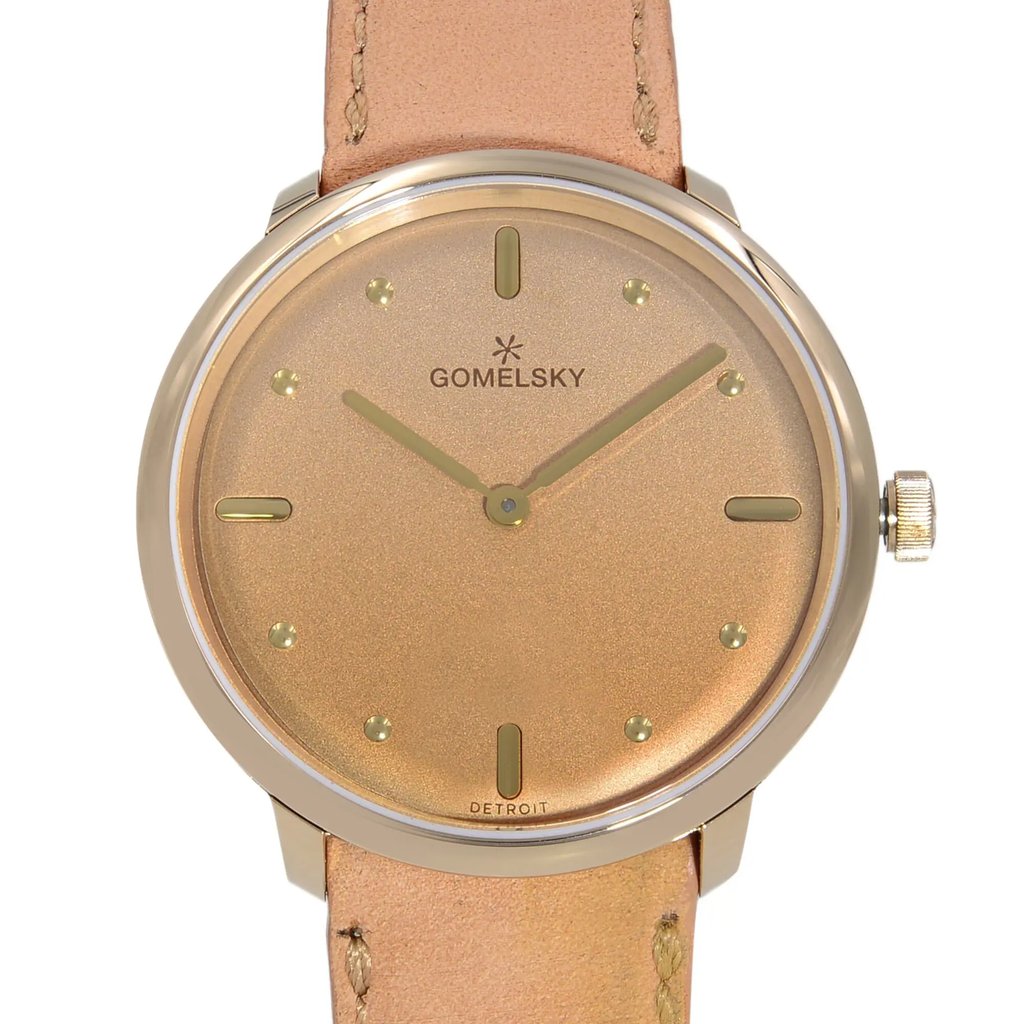 An image of a wristwatch taken from the front in a close-up shot, showcasing the watch face. The watch is unworn, with a minimalistic design featuring a gold-toned dial and matching hands. The brand name "Gomelsky" is displayed on the dial. The watch has a simple, elegant appearance with a beige leather strap visible at the top and bottom.
