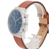 An image of a Skagen men's wristwatch displayed in a side angle view. The watch features a chronograph and date indicator, with a blue dial and silver-tone casing. It has a brown leather strap and is set on a transparent display stand. The photo is taken at a close distance, highlighting the watch's design and features.