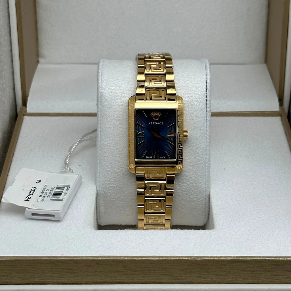 An image of a Versace women's wristwatch displayed upright on a cushioned stand. The watch features a rectangular gold case and bracelet with a geometric pattern. The watch face is dark blue with gold-tone hour markers and hands. The image is taken from a frontal angle, positioned slightly above the watch, and at a close distance, capturing detailed design elements. A tag with product information is visible to the left of the watch.