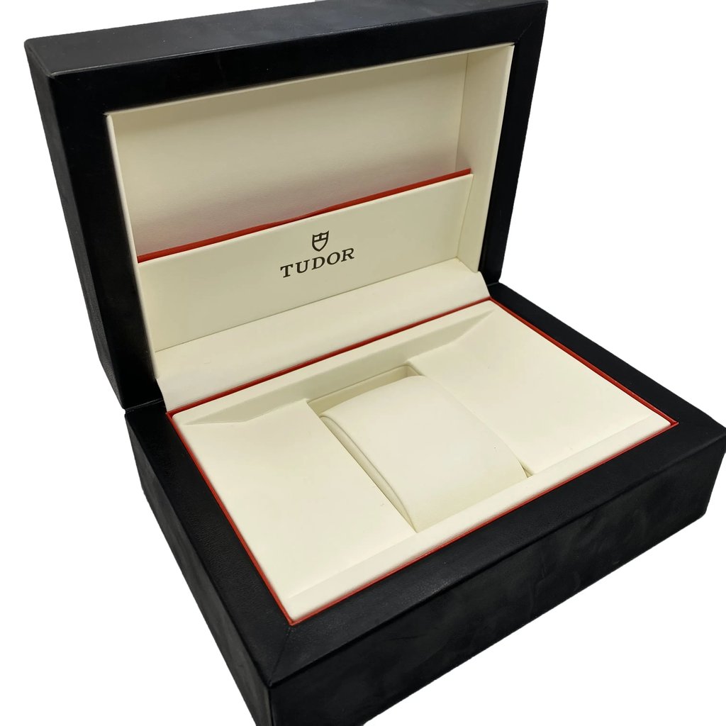 An image of an empty Tudor wristwatch box taken from a slightly elevated angle at a medium distance. The box is open, displaying a cream-colored interior with the Tudor logo on the inner lid. The exterior is black with a subtle red trim, designed to hold a men’s wristwatch.