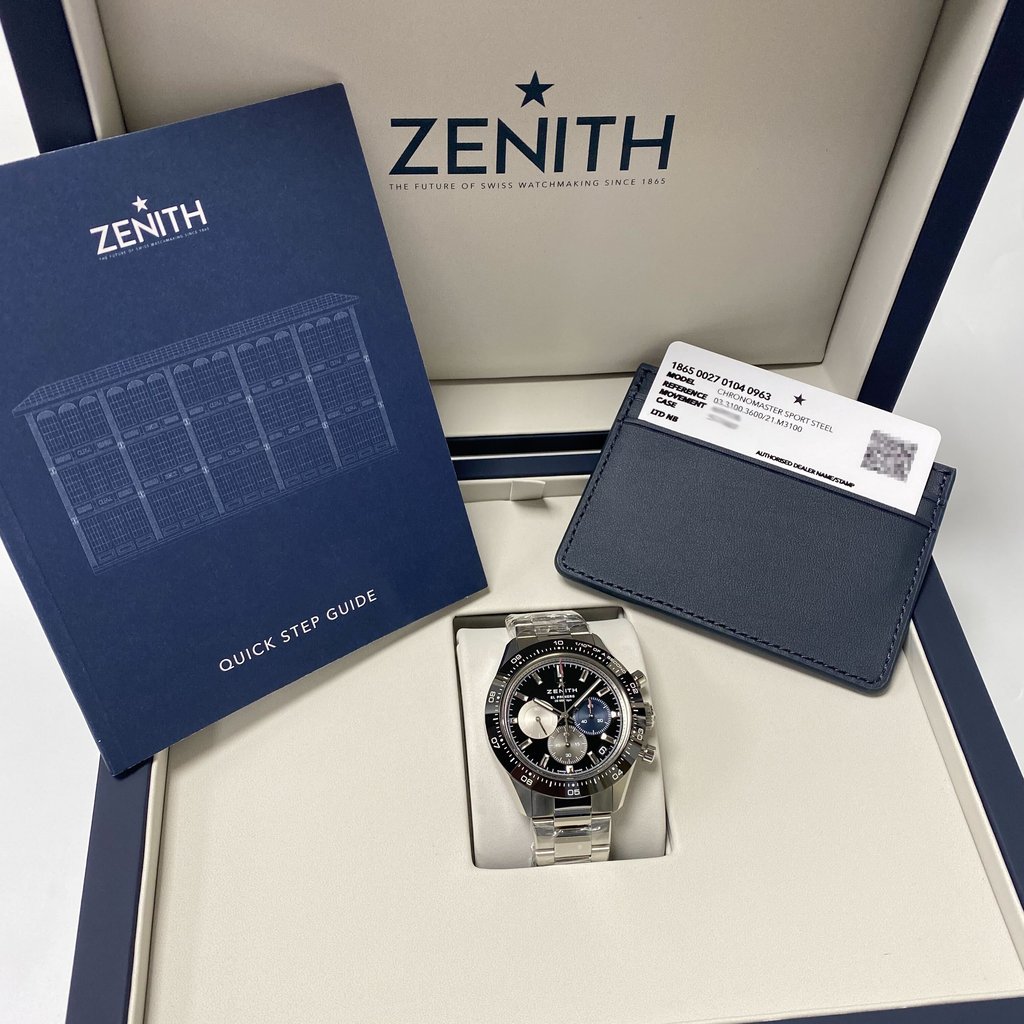 An image of an unworn wristwatch displayed inside an open box. The watch is positioned centrally, resting on a cushioned surface, with its face angled slightly upward, showcasing its dial and features. To the left, a blue "Quick Step Guide" booklet is visible, and to the right, a cardholder containing a document is placed. The box lid is open, revealing the brand's logo and tagline. The photo is taken from a top-down perspective, capturing the entire setup clearly.