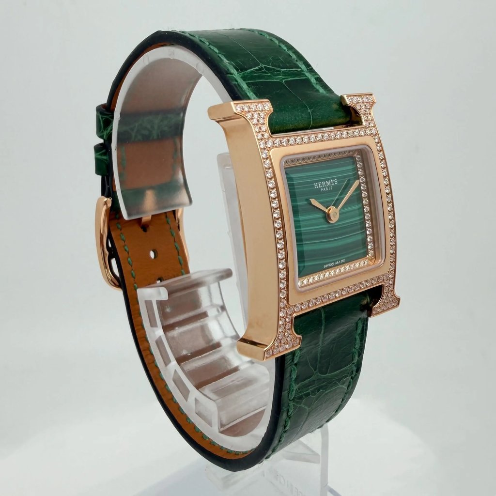 An image of a pre-owned women's HERMÈS wristwatch, positioned at a three-quarter angle. The watch features a square, gold-toned case embellished with diamonds, set against a green, textured leather strap. The watch face displays a green dial with gold hands. The image is taken at a close range, showcasing the intricate details of the watch's design.