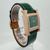 An image of a pre-owned women's HERMÈS wristwatch, positioned at a three-quarter angle. The watch features a square, gold-toned case embellished with diamonds, set against a green, textured leather strap. The watch face displays a green dial with gold hands. The image is taken at a close range, showcasing the intricate details of the watch's design.