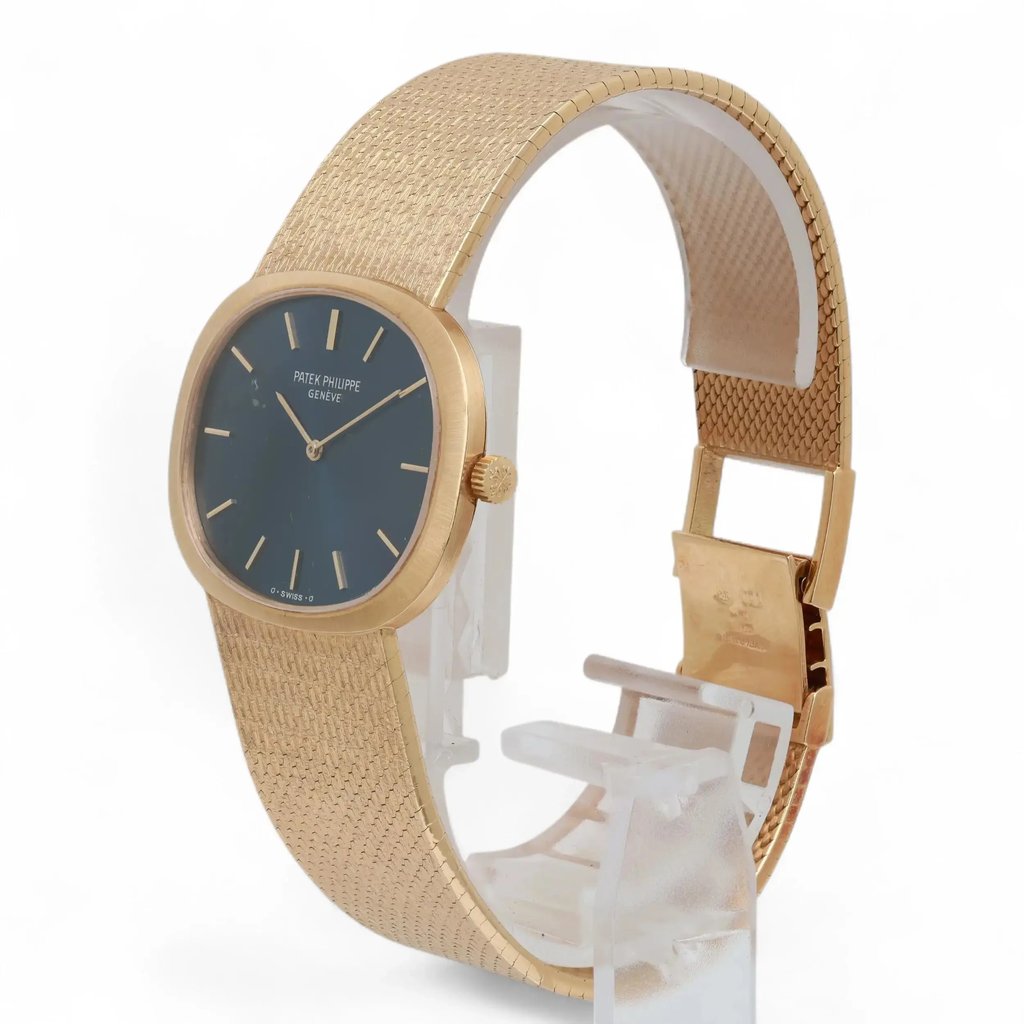 An image of a pre-owned Patek Philippe wristwatch positioned upright on a clear display stand. The watch is viewed from a slightly angled front-left position, showcasing its elegant design. The timepiece features a blue dial with yellow gold hour markers and hands, encased in a gold frame. The textured gold bracelet is partially visible, highlighting its luxurious appearance. The image is taken from a moderate distance, capturing the watch's intricate details and craftsmanship.