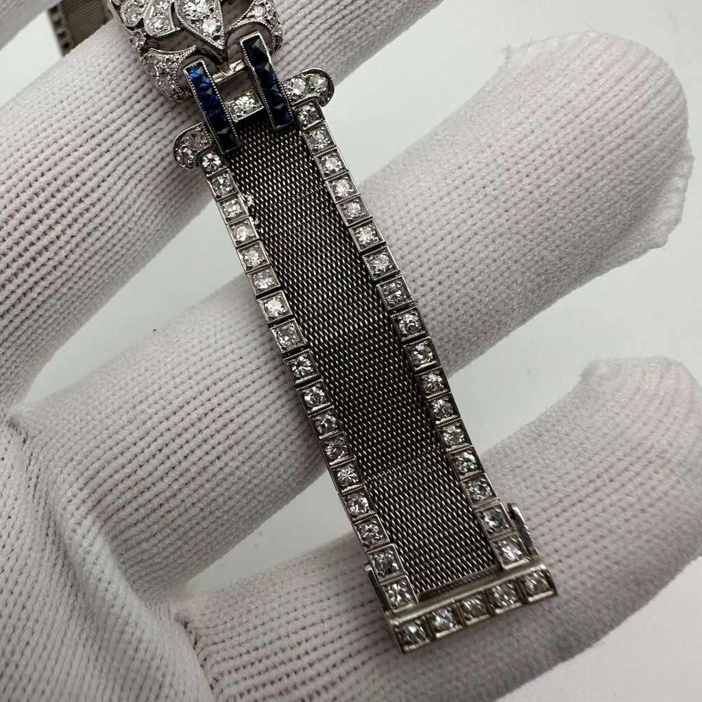 An image of a pre-owned Tissot women's wristwatch, presented at a close-up angle. The watch is positioned diagonally across the frame with the watch face towards the top left corner, partially obscured by a white-gloved hand, suggesting the watch is being held or displayed. The watch has a metallic mesh strap adorned with a double row of square-cut diamonds along its edges, leading to a decorative clasp with similar diamond embellishments. The view is focused on the strap and clasp, with the details captured from a top-down perspective.