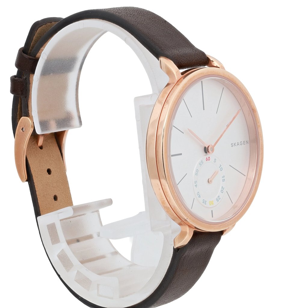 An image of a pre-owned Skagen wristwatch positioned at a three-quarter angle, showcasing both the watch face and part of the strap. The watch has a minimalist white dial with slim hour markers and a small seconds subdial. The case is a rose gold color, and the strap is dark brown leather. The watch is displayed on a white stand, and the photo is taken at a close distance to highlight the design details.