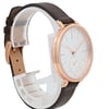 An image of a pre-owned Skagen wristwatch positioned at a three-quarter angle, showcasing both the watch face and part of the strap. The watch has a minimalist white dial with slim hour markers and a small seconds subdial. The case is a rose gold color, and the strap is dark brown leather. The watch is displayed on a white stand, and the photo is taken at a close distance to highlight the design details.