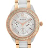 An image of a front close-up shot of a women's dress/formal Bulova wristwatch. The watch face is in clear view at a direct angle, showing a round silver sector dial with gold-tone bezel adorned with diamonds. The stainless steel band combines silver and gold-tone links, and the dial features Arabic numerals at 12, 2, 4, 6, 8, and 10 o'clock positions, with stick indexes for other hours. There are three smaller subdials indicating day, date, and GMT, and the hands are luminous. The image is taken from a short distance to capture the entire watch face and part of the band, providing a detailed view of the watch's design and features.