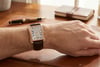 An image of a pre-owned Piaget men's wristwatch displayed on a person's wrist. The watch has a rectangular face with a rose gold case and a brown leather strap. The angle is from above, slightly to the left, showing the watch face clearly. The background includes a wooden desk surface with a pen and a notebook, giving a sense of a close and intimate setting.