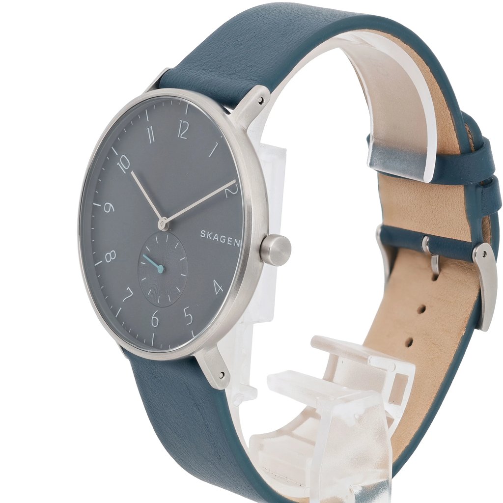 An image of a Skagen men's wristwatch positioned in a three-quarter view, captured from a slightly elevated angle. The watch features a minimalist design with a dark dial, silver hour markers, and a small seconds subdial. The blue leather strap is partially visible, and the watch is mounted on a clear display stand.