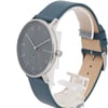An image of a Skagen men's wristwatch positioned in a three-quarter view, captured from a slightly elevated angle. The watch features a minimalist design with a dark dial, silver hour markers, and a small seconds subdial. The blue leather strap is partially visible, and the watch is mounted on a clear display stand.