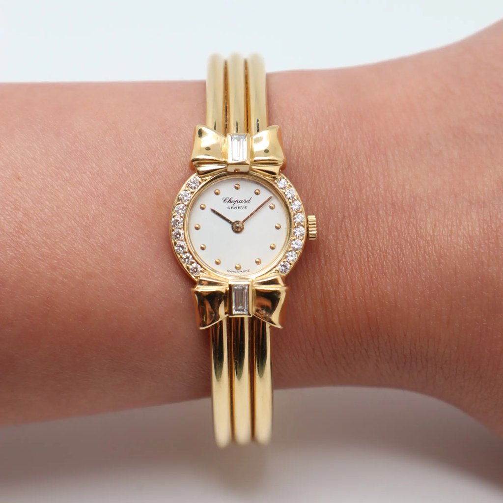 An image of a pre-owned Chopard women's wristwatch positioned on a person’s wrist. The watch is photographed from a top-down angle at a close distance. It features a yellow gold band and a round face adorned with decorative bows and diamonds around the bezel.