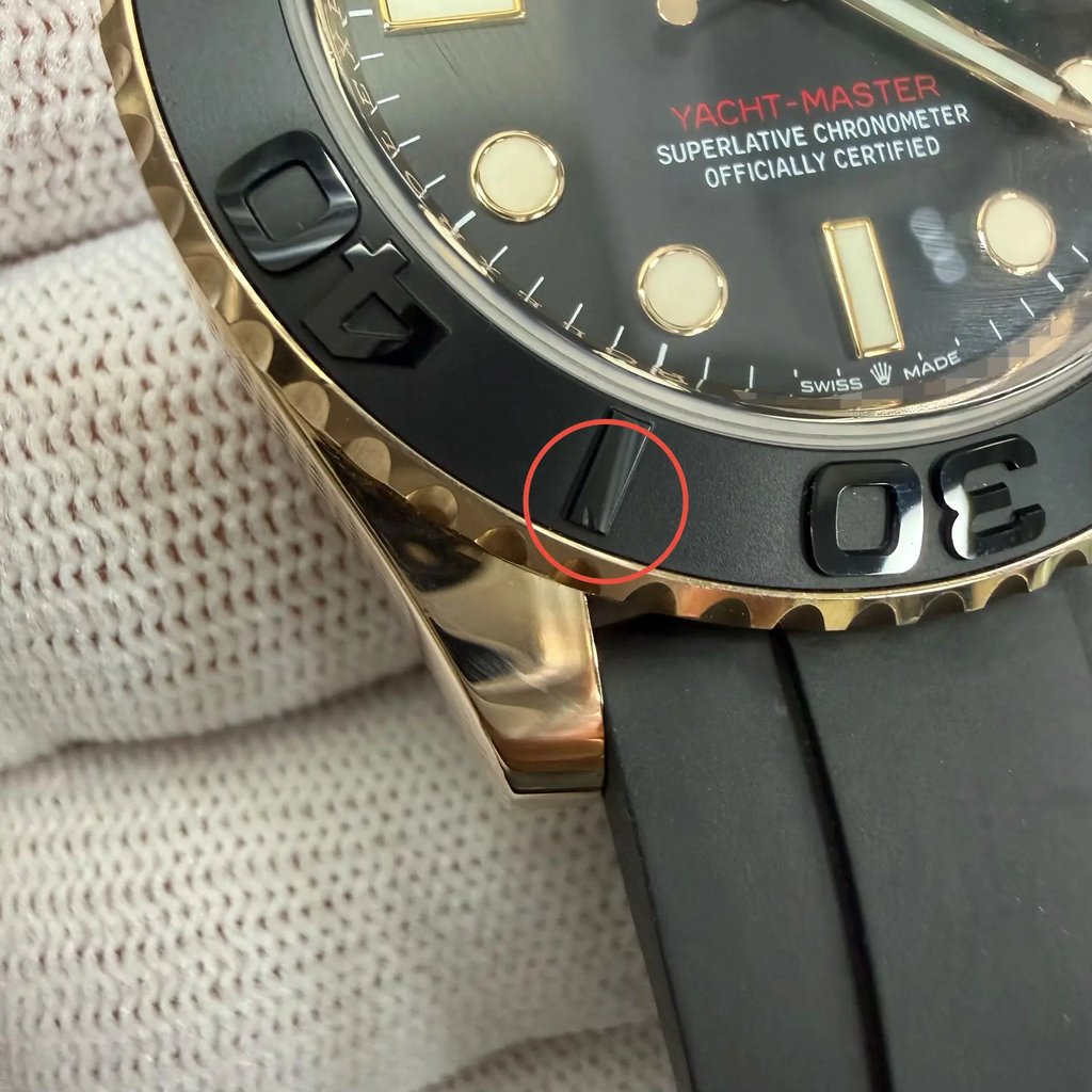 An image of a pre-owned men's Rolex wristwatch taken up close at an angle, showcasing the left side of the watch face. The watch features a black bezel with embossed numerals and a gold casing. The dial has luminous hands and a date indicator, with "Yacht-Master" in red text. The watch is held by a gloved hand, and part of the black rubber strap is visible.Minor scratch on the bezel between 30 and 40. It shows light signs of wear.