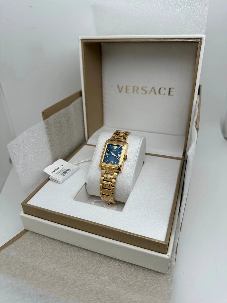 An image of a Versace women's wristwatch displayed at a slight angle, positioned centrally within an open box. The watch features a rectangular gold case and bracelet with a blue dial. The image is taken from a close distance, showcasing the elegant design and intricate details. The box is lined with white and gold, with the brand name prominently visible on the inside lid.