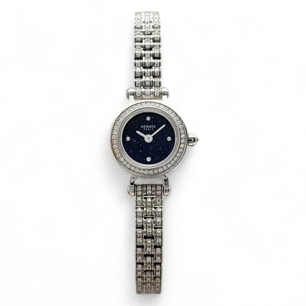 An image of a pre-owned women's Hermès Faubourg Joaillerie 16.3 mm 18K Gold Diamond Blue Dial Watch W049640WW00 taken from a top-down angle. The watch features a round face with a blue dial and minimalistic markers. The bezel and bracelet are adorned with sparkling diamonds, showcasing an elegant design. The watch is centered in the frame, photographed against a plain white background, highlighting its intricate details.