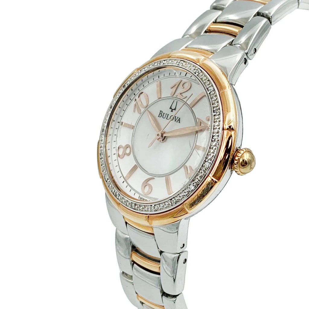 An image of a pre-owned Bulova women's wristwatch in a casual and dress/formal style. The side profile view shows the watch's rose gold-tone crown prominently. The watch has a round shape with a white dial featuring a diamond pattern and rose gold-tone bezel accented with diamonds The band is made of stainless steel with a two-tone design, combining silver and rose gold elements. The watch is photographed up close, capturing the details of the dial, bezel, and the upper part of the band.