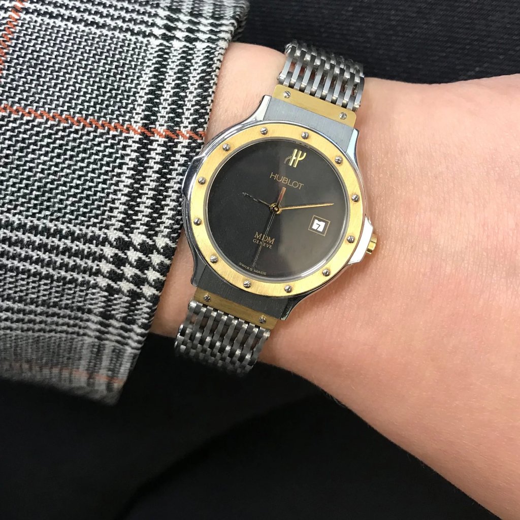 An image of a pre-owned wristwatch taken from a front full-frontal view. The watch is positioned on a wrist, showing its round face with a gold bezel and a metallic strap. The angle is direct, capturing the watch face clearly, with details such as the brand name visible. The distance is close enough to showcase the intricate design and condition of the timepiece. The wrist is adorned with a patterned fabric sleeve.