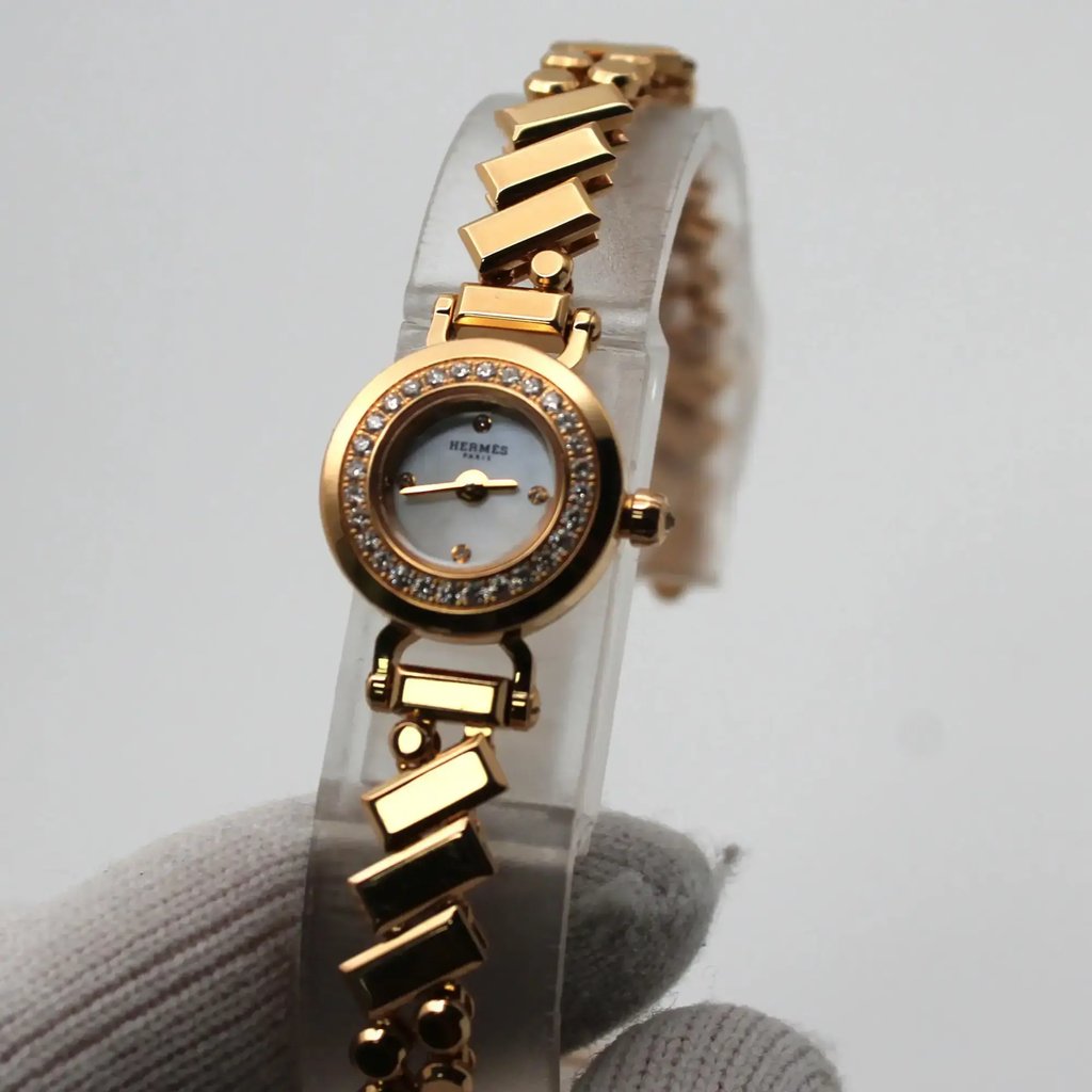 An image of a pre-owned women's HERMÈS wristwatch displayed at a slight angle, positioned vertically on a clear stand. The watch features a round face with a bezel adorned with small diamonds. The rose gold linked bracelet has a unique geometric design. The watch is held at a close distance by a gloved hand, showcasing its elegant details.