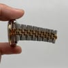 An image of a pre-owned Rolex unisex wristwatch held between fingers, viewed from the side. The watch features a gold and steel metal bracelet with a prominent date indicator. The angle shows the watch's profile and band detail, with the camera positioned close to capture the intricate design.