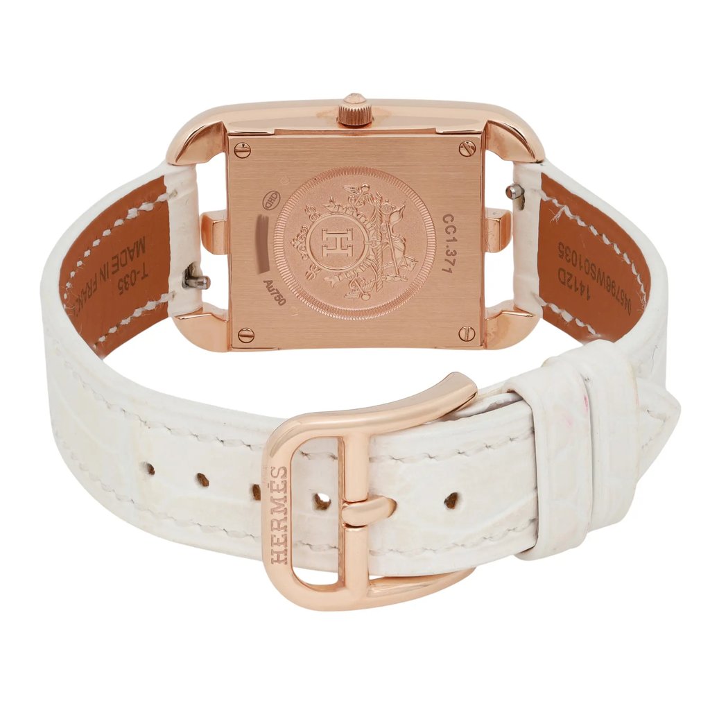 An image of a pre-owned women's HERMÈS Cod Joaillier wristwatch, viewed from the back at a slight angle. The focus is on the rectangular, rose gold-toned case, featuring intricate engravings. The white leather strap is visible, with the HERMÈS logo embossed on the clasp. The watch is positioned centrally and captured from a close distance, highlighting its elegant design and craftsmanship.