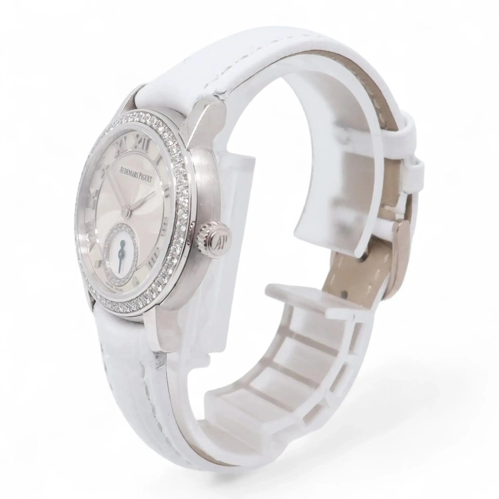 An image of a pre-owned Audemars Piguet women's wristwatch, viewed from a slightly elevated angle, showcasing the front and side. The watch features a round face with a diamond-studded bezel and a white leather strap. The watch is positioned to highlight its elegant design details, with the crown on the right side visible. The photograph is taken at a close distance, emphasizing the luxurious elements of the timepiece.